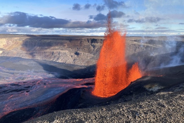 Lava fountain erupting in a volcanic crater under blue sky.
