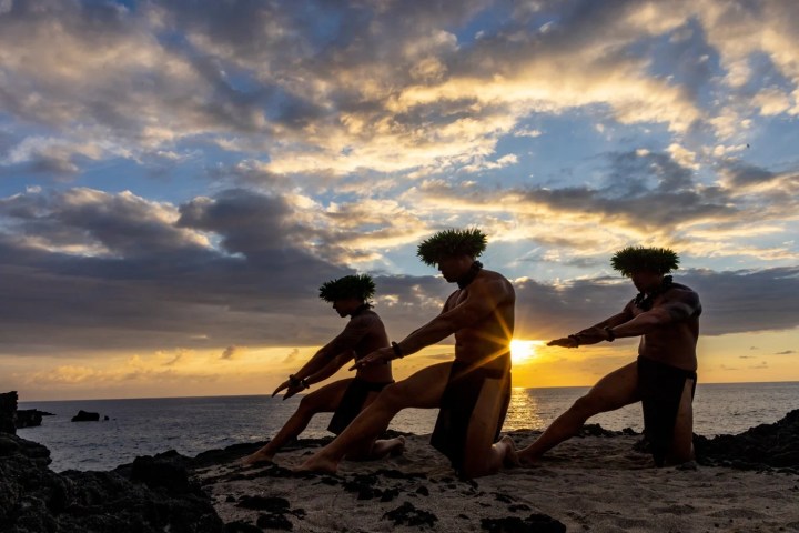 Three dancers in traditional attire on a beach at sunset.