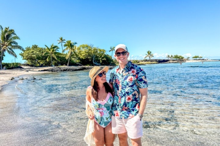 Couple in floral outfits standing on a beach with palm trees and clear blue sky.