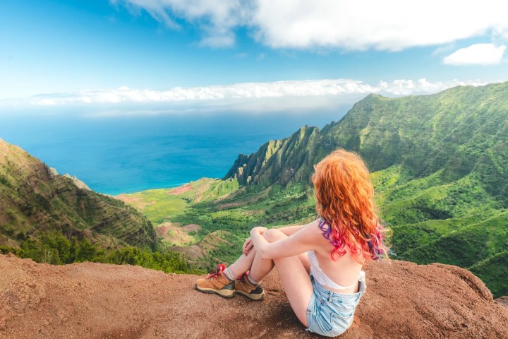 Person with colorful hair sitting on cliff, overlooking lush valley and ocean.