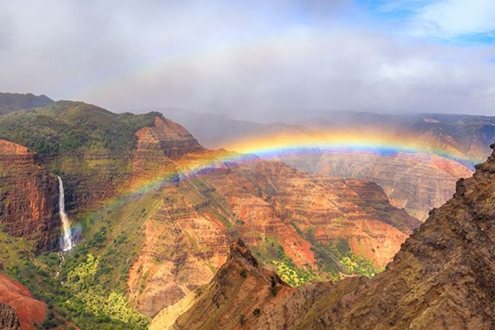 Rainbow over a vibrant canyon with a waterfall and cloudy sky.